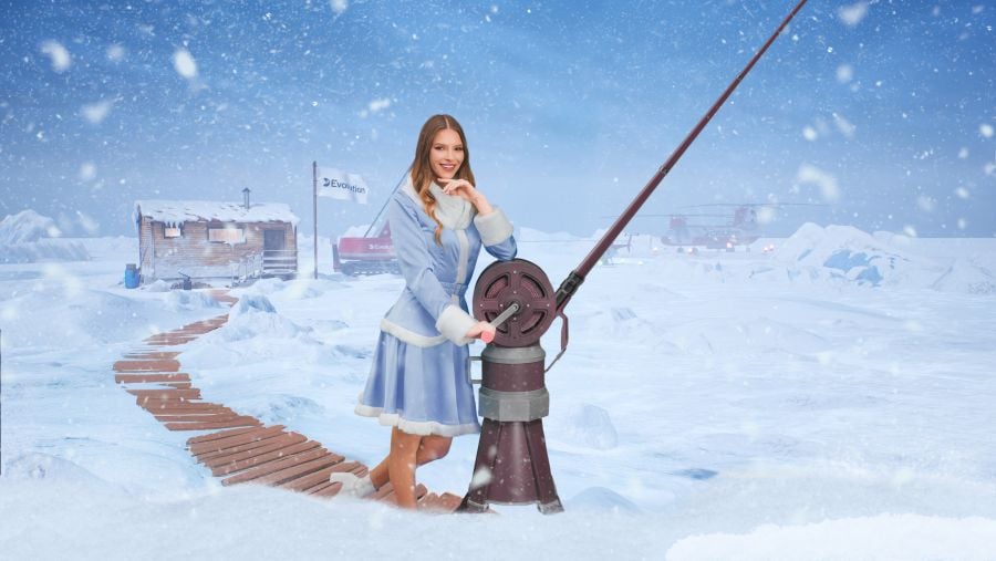 Female live game show host in a blue fur-trimmed winter coat standing beside a large fishing reel on a snowy Arctic set with a wooden path, cabin, and Evolution-branded flags in the background. - foxybingo